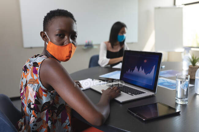 Retrato de mulher afro-americana usando máscara facial usando laptop na sala de reuniões do escritório moderno. bloqueio de quarentena por distanciamento social durante a pandemia do coronavírus — Fotografia de Stock