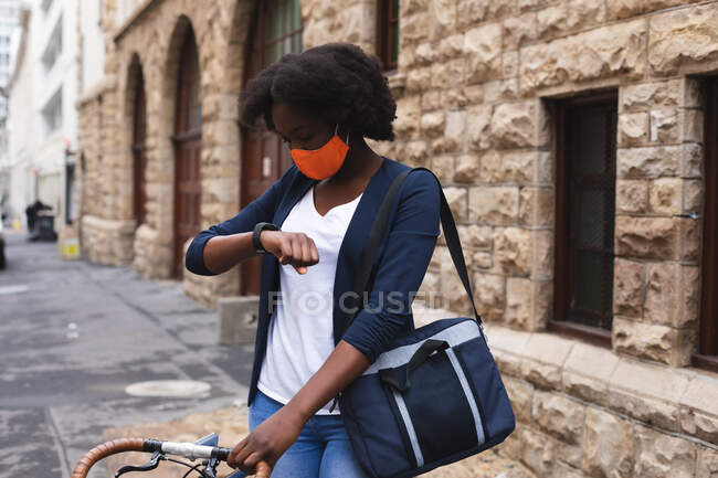 Mulher afro-americana usando máscara facial na rua de pé e verificando seu smartwatch. fora e sobre na cidade durante covid 19 coronavirus pandemia. — Fotografia de Stock