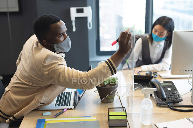 Homem afro-americano usando máscara facial escrevendo em placa de vidro no escritório moderno. higiene e distanciamento social no local de trabalho durante coronavírus covid 19 pandemia. — Fotografia de Stock