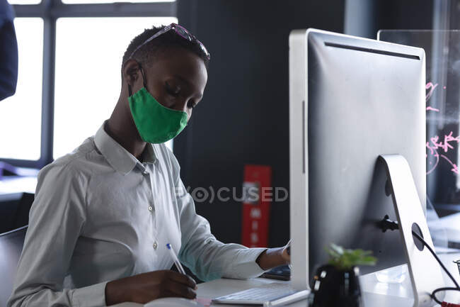 Африканская американка в маске работает, сидя на столе в современном офисе. социальная изоляция от карантина во время пандемии коронавируса — стоковое фото