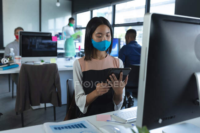 Азійка, одягнена в маску обличчя, користується цифровим планшетом, сидячи на столі в сучасному офісі. гігієна і соціальна дистанція на робочому місці під час коронавірусу covid 19 пандемії. — стокове фото
