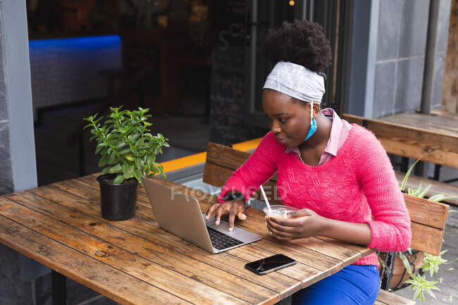 Africano americano mulher sentado em um café usando um laptop e comer uma salada fora e sobre na cidade durante covid 19 coronavirus pandemia. — Fotografia de Stock