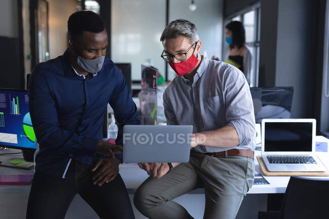 Разнообразные коллеги-мужчины в масках для лица используют ноутбук, сидя над своим столом в современном офисе. социальная изоляция от карантина во время пандемии коронавируса — стоковое фото