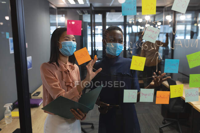 Різні жінки-колеги, одягнені в маски для мозкового штурму в офісі. Обговорюючи записки по скляній стіні в сучасному офісі. гігієна на робочому місці під час пандемії коронавірусу. — стокове фото