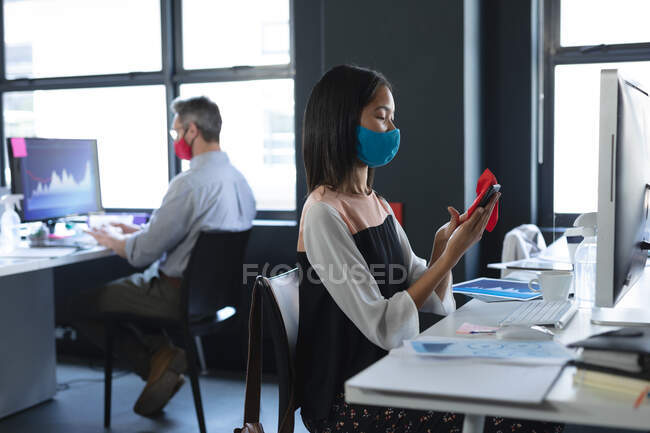 Mulher asiática usando máscara facial limpando seu smartphone enquanto sentada em sua mesa no escritório moderno. higiene e distanciamento social no local de trabalho durante coronavírus covid 19 pandemia. — Fotografia de Stock