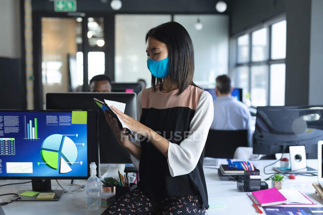 Азійка, одягнена в маску, прибирає в сучасному офісі свій цифровий планшет. гігієна і соціальна дистанція на робочому місці під час коронавірусу covid 19 пандемії. — стокове фото