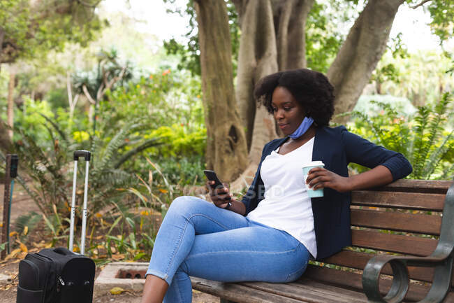 Donna afroamericana con maschera in strada seduta su una panchina, che beve una tazza di caffe '. in giro per la città durante covid 19 coronavirus pandemia. — Foto stock