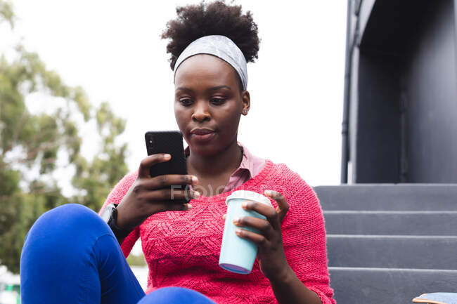 Femme afro-américaine utilisant un smartphone et tenant la tasse dans la rue dans la ville pendant la pandémie de coronavirus covid 19. — Photo de stock