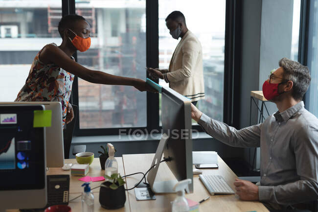 Різні чоловічі та жіночі колеги носять маски для обличчя в офісі. жінка передає документ чоловікові, який сидить за столом. карантин соціального дистанціювання під час пандемії коронавірусу — стокове фото