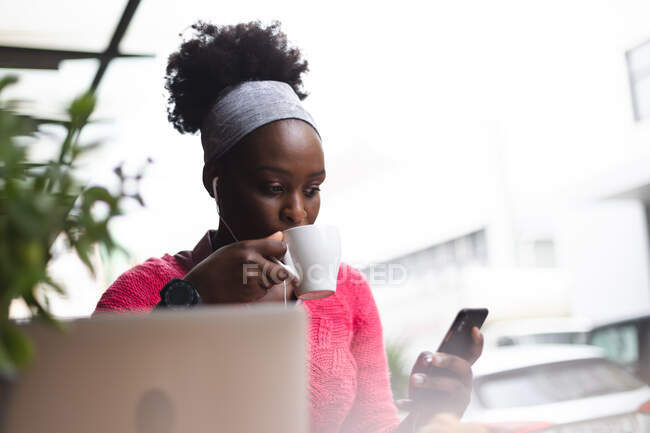 Mujer afroamericana sentada en un café usando un smartphone, bebiendo una taza de café y escuchando música. fuera de la ciudad durante la pandemia de coronavirus covid 19. - foto de stock