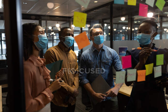 Diversos colegas de escritório usando máscaras faciais discutindo sobre notas de memorando sobre placa de vidro no escritório moderno. bloqueio de quarentena por distanciamento social durante a pandemia do coronavírus — Fotografia de Stock
