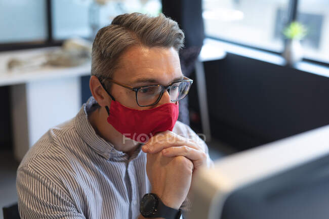 Белый мужчина в маске смотрит на экран компьютера в офисе. социальная изоляция от карантина во время пандемии коронавируса. — стоковое фото