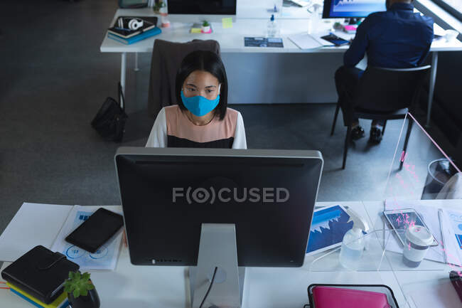 Vista aérea de una mujer asiática que usa mascarilla usando computadora mientras está sentada en su escritorio en la oficina moderna. higiene y distanciamiento social en el lugar de trabajo durante la pandemia de coronavirus covid 19. - foto de stock