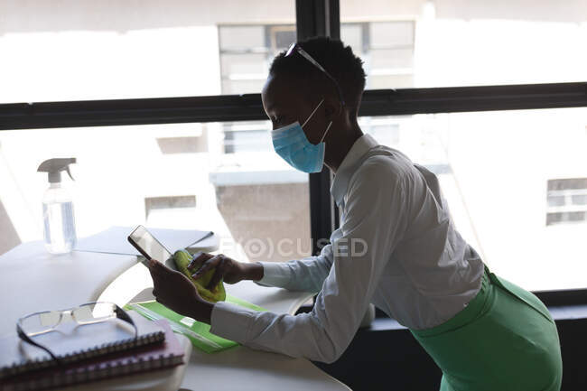 Африканська американка, одягнена в маску обличчя, чистить свою цифрову таблетку тканиною в сучасному офісі. Відстань до карантину під час пандемії коронавірусу — стокове фото