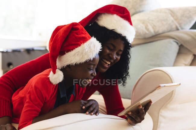 Afroamericana madre e hija teniendo video chat en tableta usando sombreros de santa durante la Navidad en casa distanciamiento social durante el bloqueo de cuarentena covid 19 coronavirus. - foto de stock
