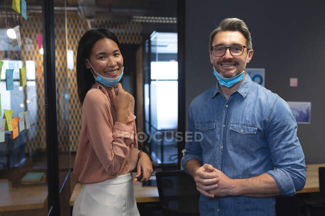 Portrait of caucasian man and asian woman with face masks around their neck smiling while standing in modern office. social distancing quarantine lockdown during coronavirus pandemic — Stock Photo
