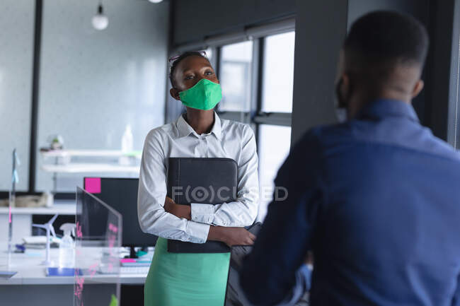 Afroamerikanerin mit Gesichtsmaske im Gespräch mit ihrem Bürokollegen im modernen Büro. Soziale Distanzierung von Quarantäne während der Coronavirus-Pandemie — Stockfoto