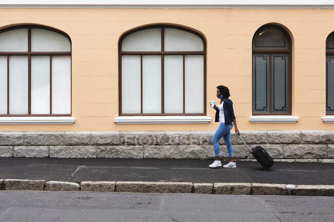 Mulher afro-americana usando máscara facial na rua bebendo uma xícara de café e carregando uma mala. fora e sobre na cidade durante covid 19 coronavirus pandemia. — Fotografia de Stock