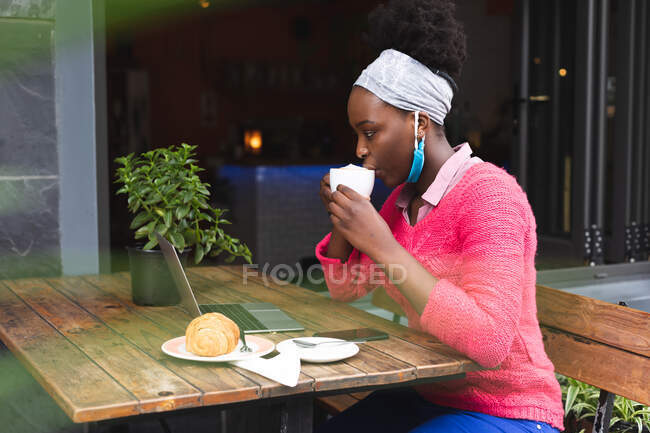 Mulher afro-americana sentada em um café usando um laptop, bebendo uma xícara de café e comendo um croissant. fora e sobre na cidade durante covid 19 coronavirus pandemia. — Fotografia de Stock