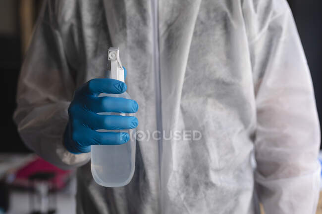 Средняя секция медицинского работника в защитной одежде с бутылкой дезинфицирующего средства в современном офисе. социальная изоляция от карантина во время пандемии коронавируса — стоковое фото