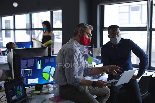 Divers collègues masculins utilisant un ordinateur portable tout en étant assis sur leur bureau au bureau moderne. isolement social mise en quarantaine pendant une pandémie de coronavirus — Photo de stock
