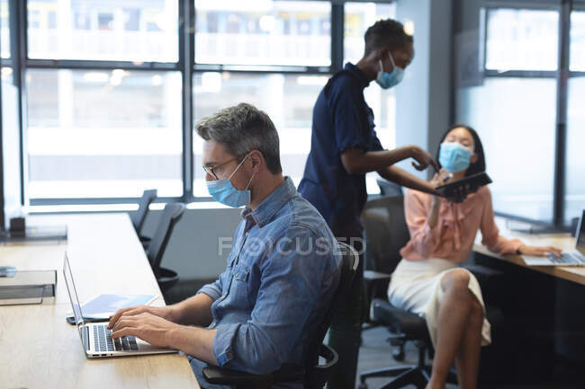 Кавказький чоловік одягнений в маску обличчя за допомогою ноутбука, в той час як афро-американська жінка і азійка, одягнені в маски для обличчя обговорюють цифрові таблетки в сучасному офісі. Відстань між людьми під час пандемії — стокове фото