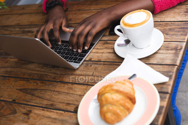 Africano americano mulher sentado em um café usando um laptop, beber uma xícara de café e comer um croissant fora e sobre na cidade durante covid 19 coronavirus pandemia. — Fotografia de Stock