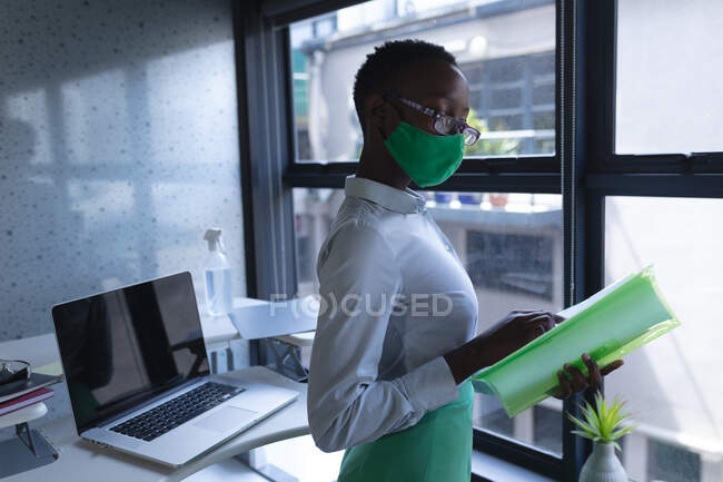 Африканская американка в маске для лица читает документы в современном офисе. гигиена и социальное дистанцирование на рабочем месте во время пандемии коронавируса 19. — стоковое фото