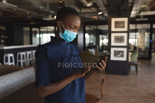 Mujer afroamericana con máscara facial usando tableta digital en la oficina moderna. distanciamiento social bloqueo de cuarentena durante la pandemia de coronavirus - foto de stock