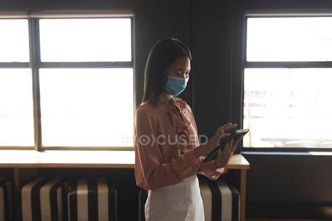 Азійка, одягнена в маску обличчя, користується цифровим планшетом у сучасному офісі. Відстань до карантину під час пандемії коронавірусу — стокове фото