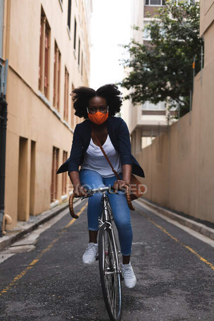 African american woman wearing face mask in street riding a bicycle out and about in the city during covid 19 coronavirus pandemic. — Stock Photo