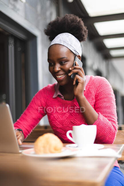 Africano americano mulher sentado em um café usando um laptop e falando em um telefone fora e sobre na cidade durante covid 19 coronavirus pandemia. — Fotografia de Stock