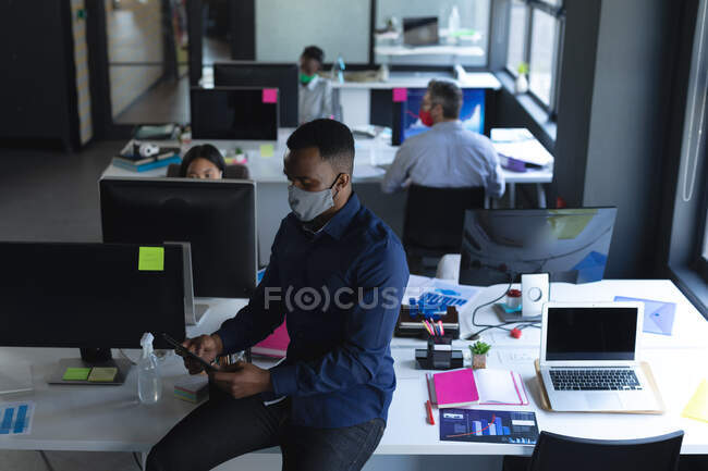 Homem afro-americano usando máscara facial usando tablet no escritório. bloqueio de quarentena por distanciamento social durante a pandemia do coronavírus. — Fotografia de Stock