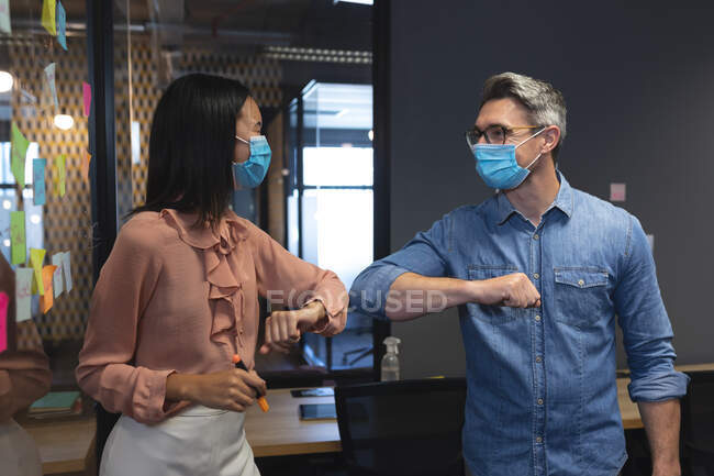Kaukasische Männer und asiatische Frauen mit Gesichtsmasken begrüßen sich im modernen Büro, indem sie die Ellbogen berühren. Soziale Distanzierung von Quarantäne während der Coronavirus-Pandemie — Stockfoto