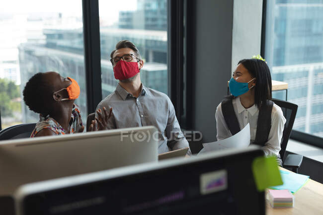 Diversos colegas con máscaras faciales riendo juntos en la oficina moderna. higiene y distanciamiento social en el lugar de trabajo durante la pandemia de coronavirus covid 19. - foto de stock