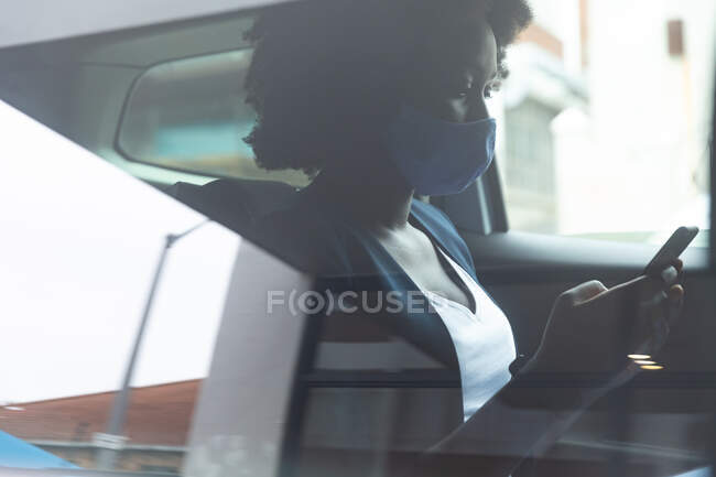 Afroamerikanerin mit Gesichtsmaske im Auto unterwegs mit Smartphone während der 19-jährigen Coronavirus-Pandemie. — Stockfoto