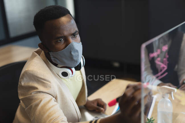 African american man wearing face mask writing on glass board at modern office. social distancing quarantine lockdown during coronavirus pandemic — Stock Photo