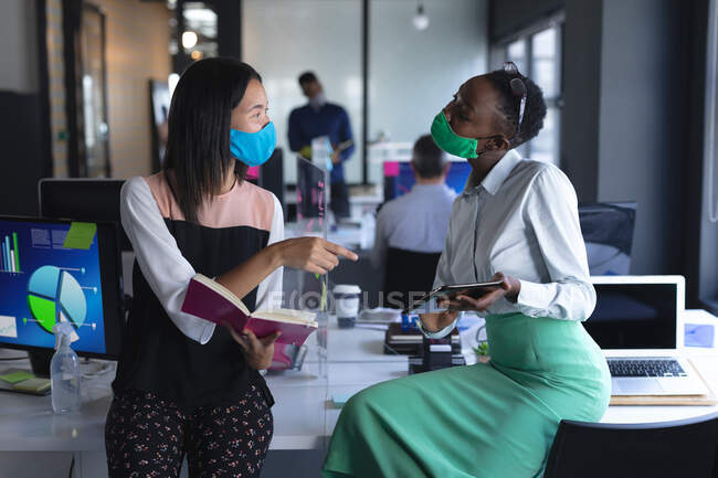 Femme asiatique avec livre et femme afro-américaine avec tablette numérique parlant entre eux au bureau moderne. hygiène et distance sociale sur le lieu de travail lors d'une pandémie de coronavirus covid 19. — Photo de stock