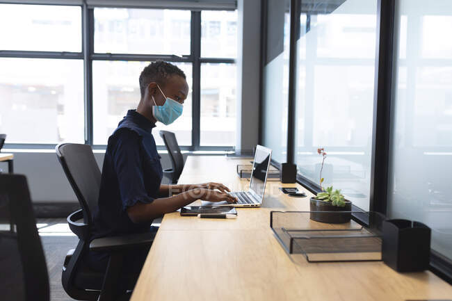 Mujer afroamericana usando mascarilla usando laptop mientras está sentada en su escritorio en la oficina moderna. distanciamiento social bloqueo de cuarentena durante la pandemia de coronavirus - foto de stock