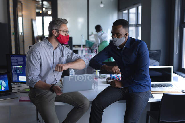 Коллеги-мужчины в масках приветствуют друг друга прикосновением к локтям в современном офисе. социальная изоляция от карантина во время пандемии — стоковое фото