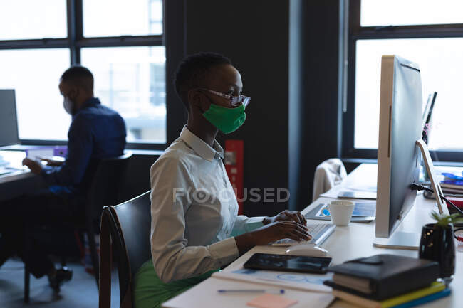 Африканська жінка-американка, яка носить маску обличчя за допомогою комп'ютера, сидячи на столі в сучасному офісі. Відстань до карантину під час пандемії коронавірусу — стокове фото