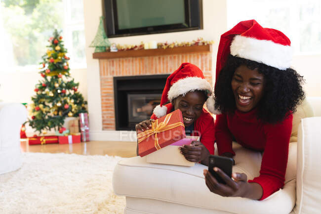Mãe e filha afro-americana conversando por vídeo no tablet. vestindo chapéus de Papai Noel durante o Natal em casa, menina está abrindo um presente. distanciamento social durante o bloqueio de quarentena do coronavírus covid 19 — Fotografia de Stock