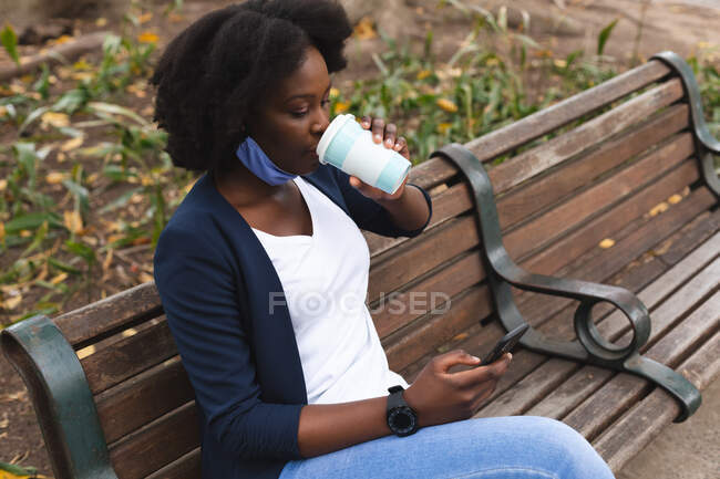 Afroamerikanerin mit Gesichtsmaske sitzt auf der Straße auf einer Bank und trinkt während der 19. Coronavirus-Pandemie in der Stadt eine Tasse Kaffee. — Stockfoto