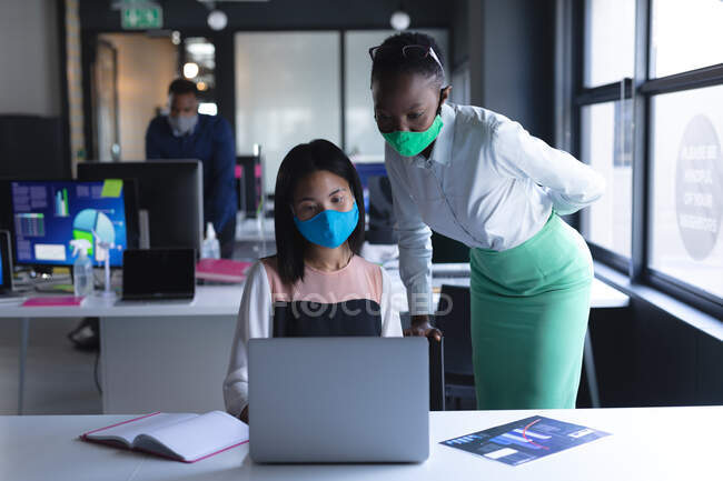 Diversos colegas do sexo feminino usando máscaras faciais usando laptop no escritório moderno. bloqueio de quarentena por distanciamento social durante a pandemia do coronavírus — Fotografia de Stock