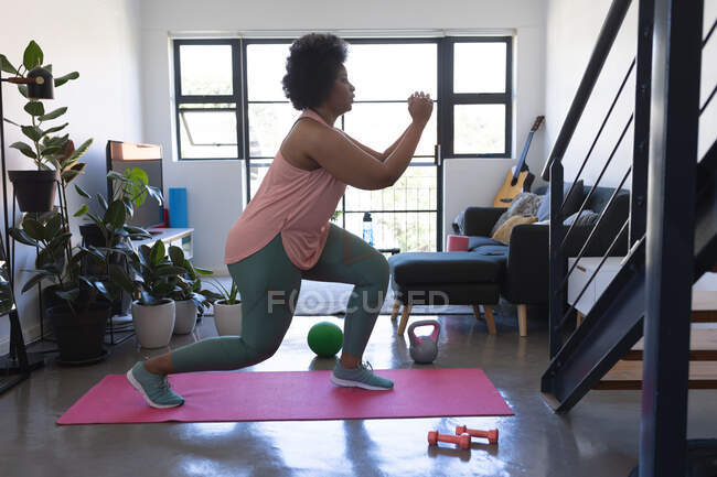 Mujer afroamericana de pie en la esterilla de ejercicio haciendo ejercicio. autoaislamiento fitness en casa durante coronavirus covid 19 pandemia. - foto de stock