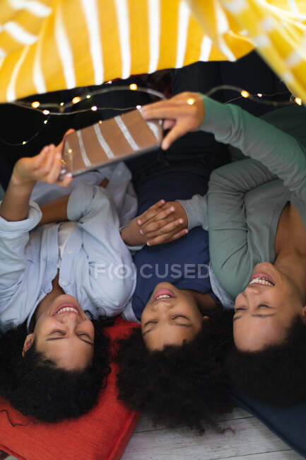 Happy mixed race lesbian couple and daughter lying down using smartphone. self isolation quality family time at home together during coronavirus covid 19 pandemic. — Stock Photo