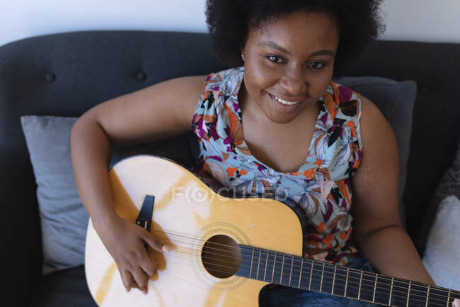 Mulher afro-americana sorridente sentada no sofá a tocar guitarra acústica. auto isolamento hobby tempo música em casa durante coronavírus covid 19 pandemia. — Fotografia de Stock