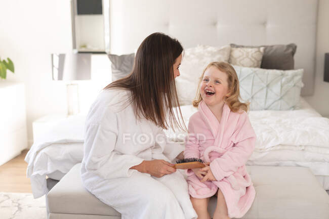 Mujer caucásica y su hija se divierten en el dormitorio. Mamá está cepillando el pelo de su hija. disfrutando de tiempo de calidad en casa durante coronavirus covid 19 pandemia de bloqueo. - foto de stock