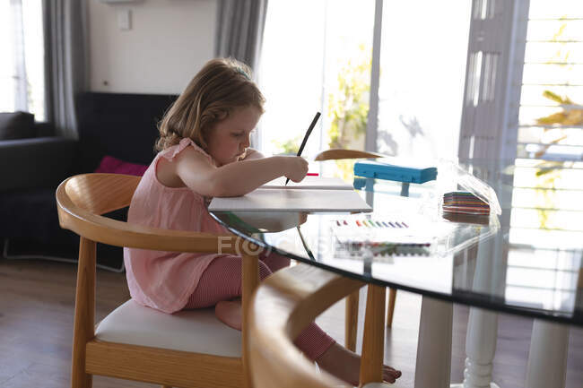 Chica caucásica sentada a la mesa dibujando en un cuaderno. disfrutando de tiempo de calidad en casa durante coronavirus covid 19 pandemia de bloqueo. - foto de stock