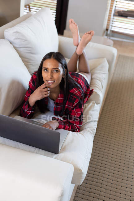 Змішана расова жінка лежить на дивані вдома, використовуючи ноутбук і посміхаючись. самоізоляція під час пандемії коронавірусу 19 . — стокове фото